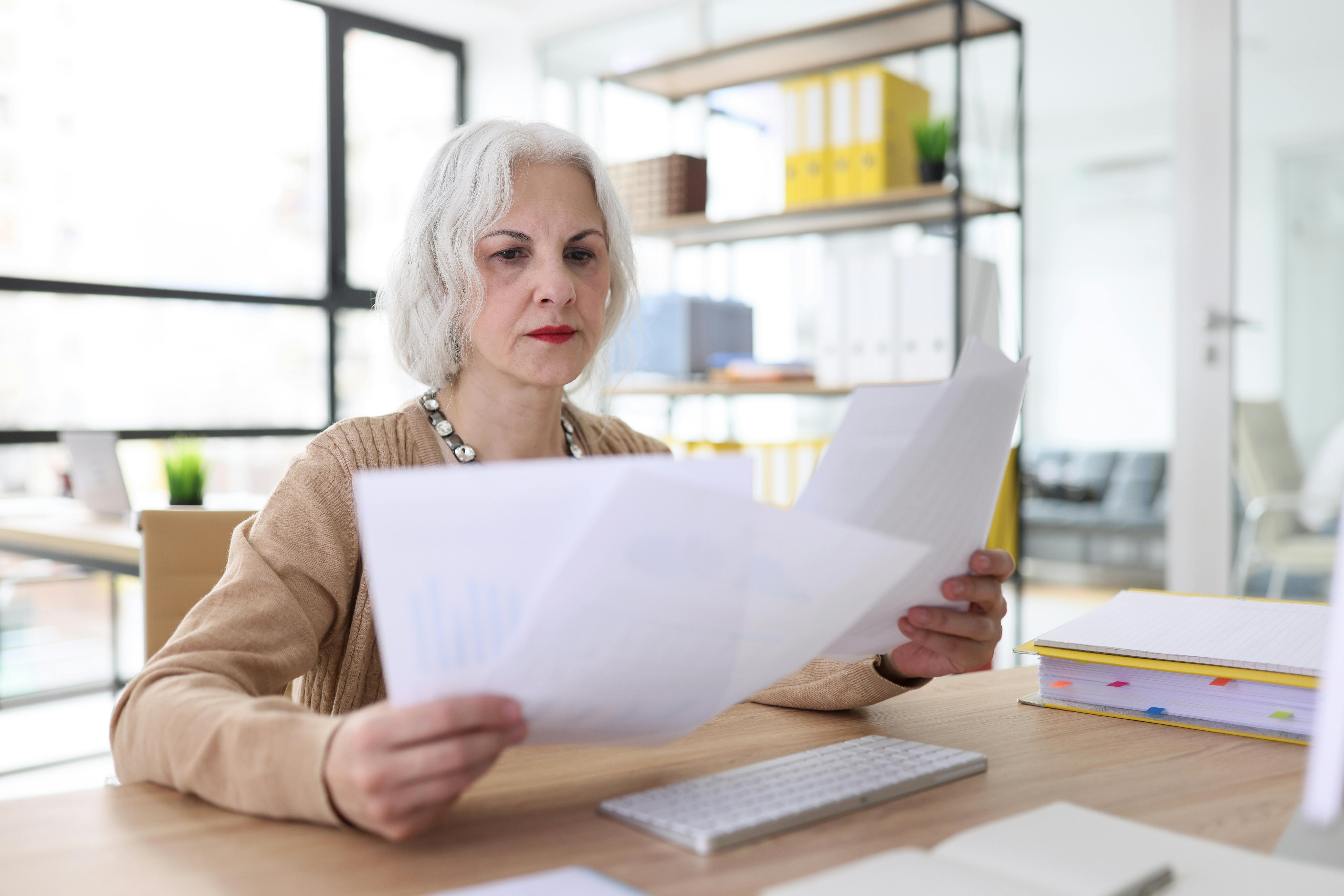 Midaldrende kvinde sidder ved sit skrivebord og ser granskende på nogle papirer, som hun holder frem foran sig.foran sig, som hun ser granskende på. H