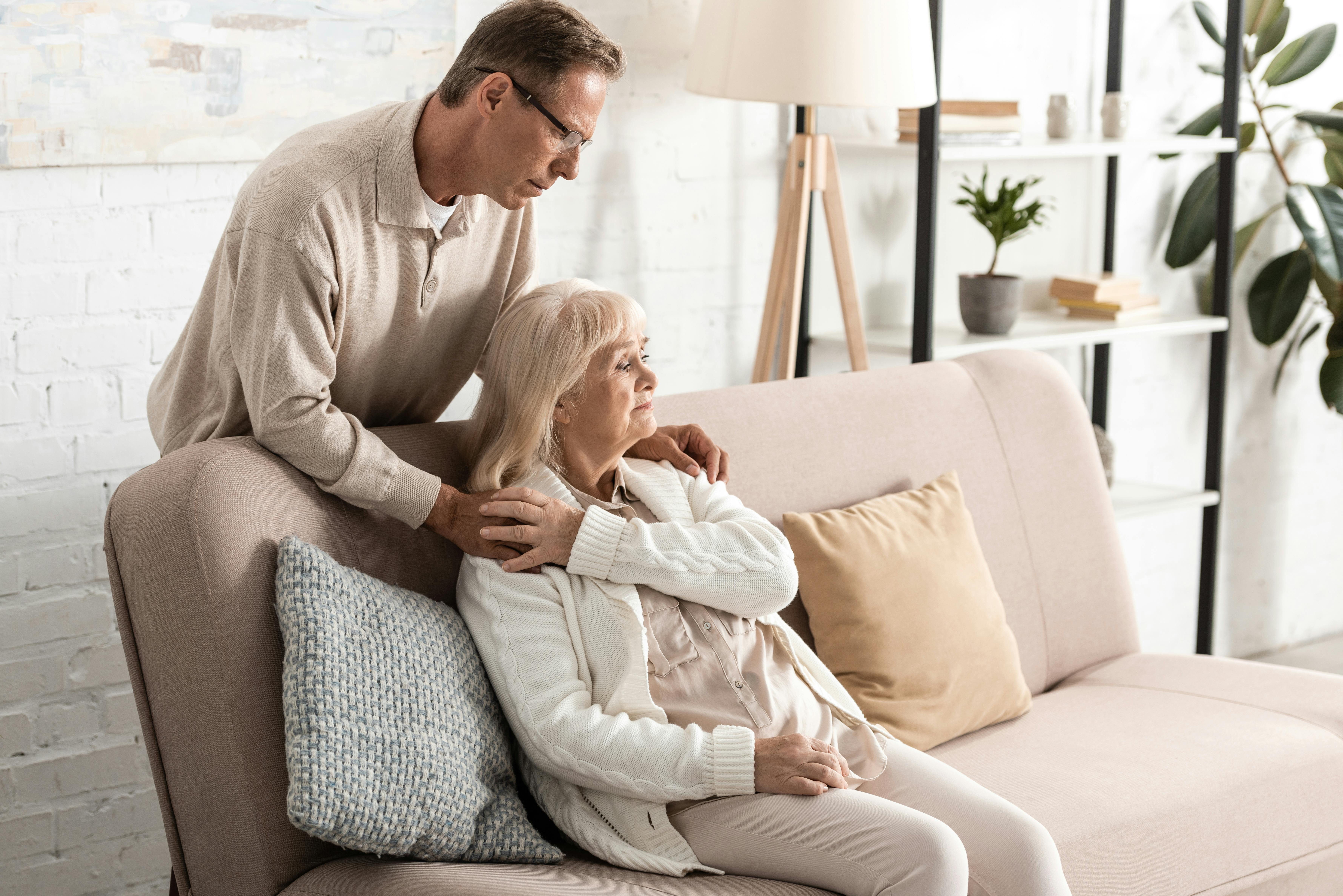 En midaldrende kvinde sidder i sin sofa og bagved står hendes mand og lægge hænderne på hendes skuldre. De ser begge alvorlige ud, og hun klapper hans ene hånd.