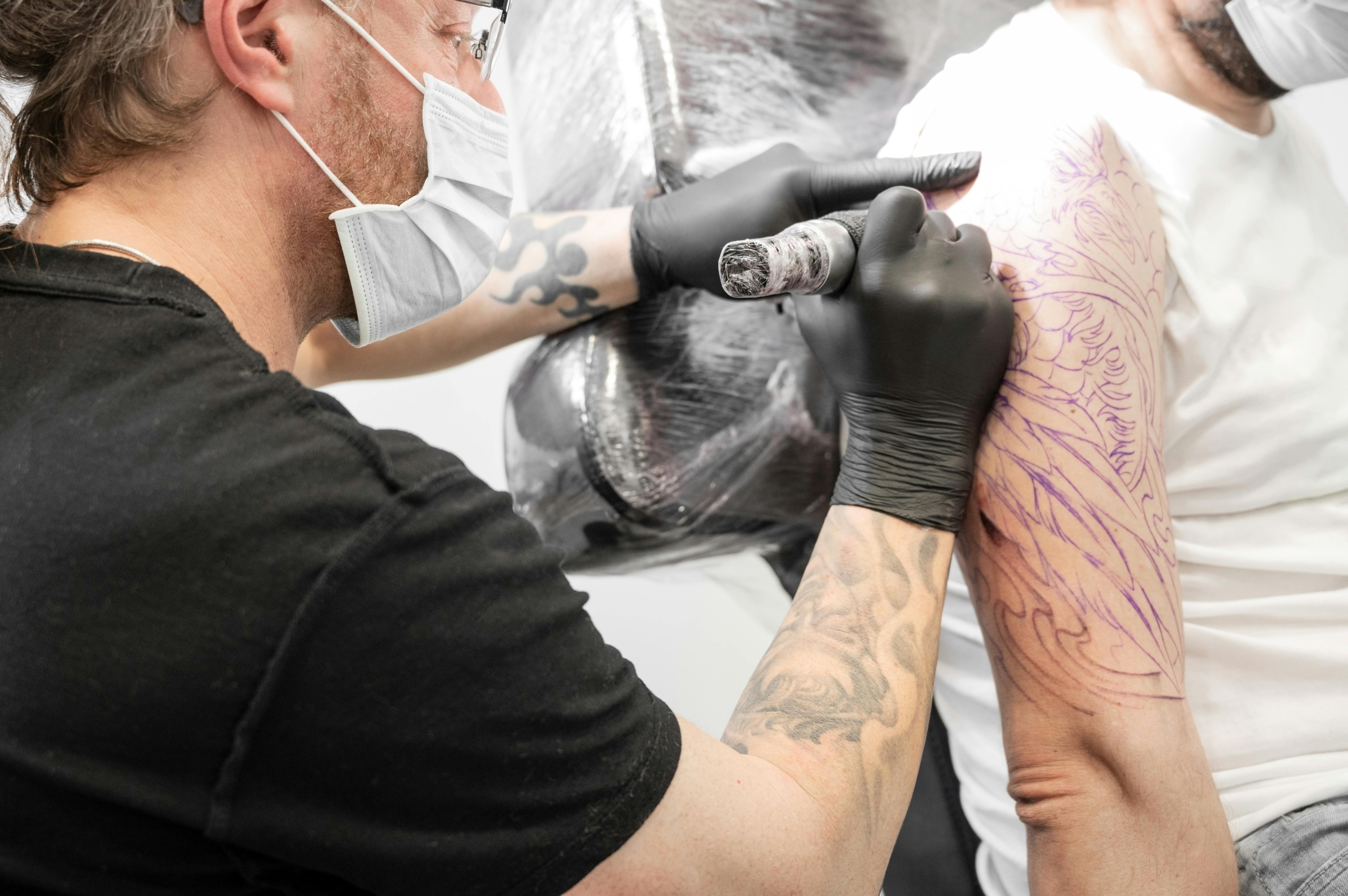 En mand er ved at tatovere en mandlig kundes overarm. 