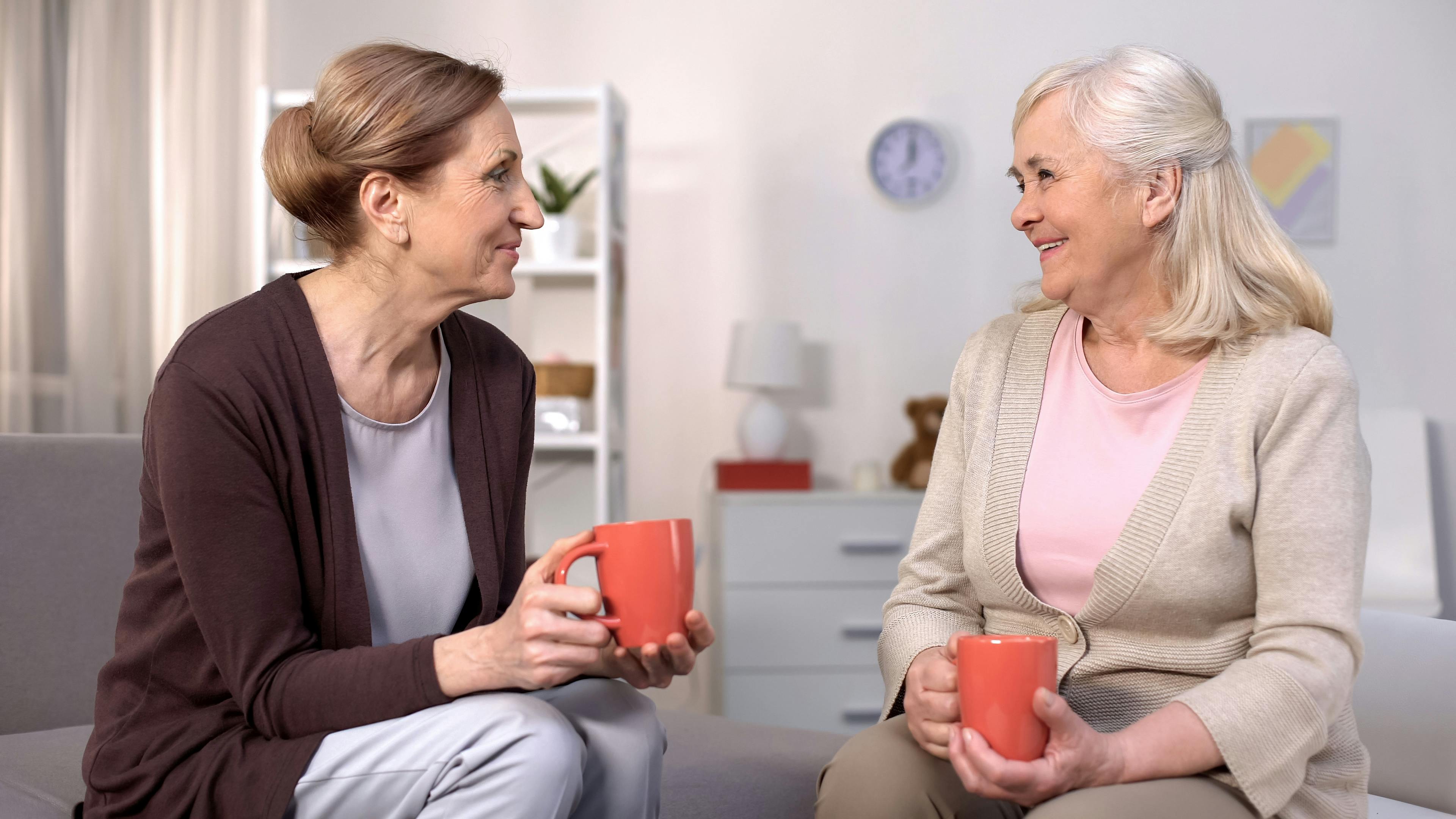 To midaldrende veninder sidder med hver sit krus kaffe og ser smilende på hinanden.