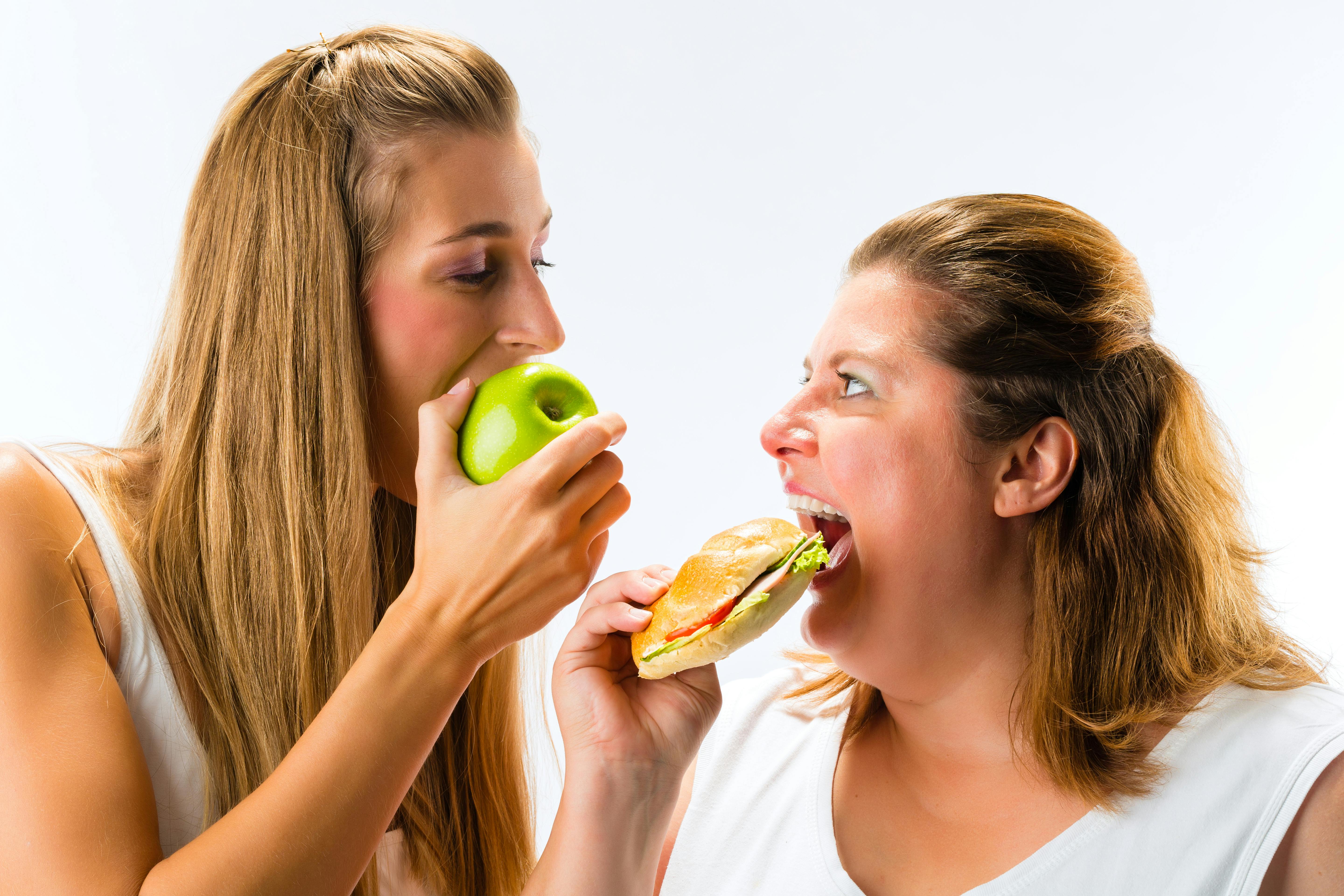 En tynd kvinde spiser et æble, mens en overvægtig kvinde spiser en sandwich. De ser hinanden i øjnene.