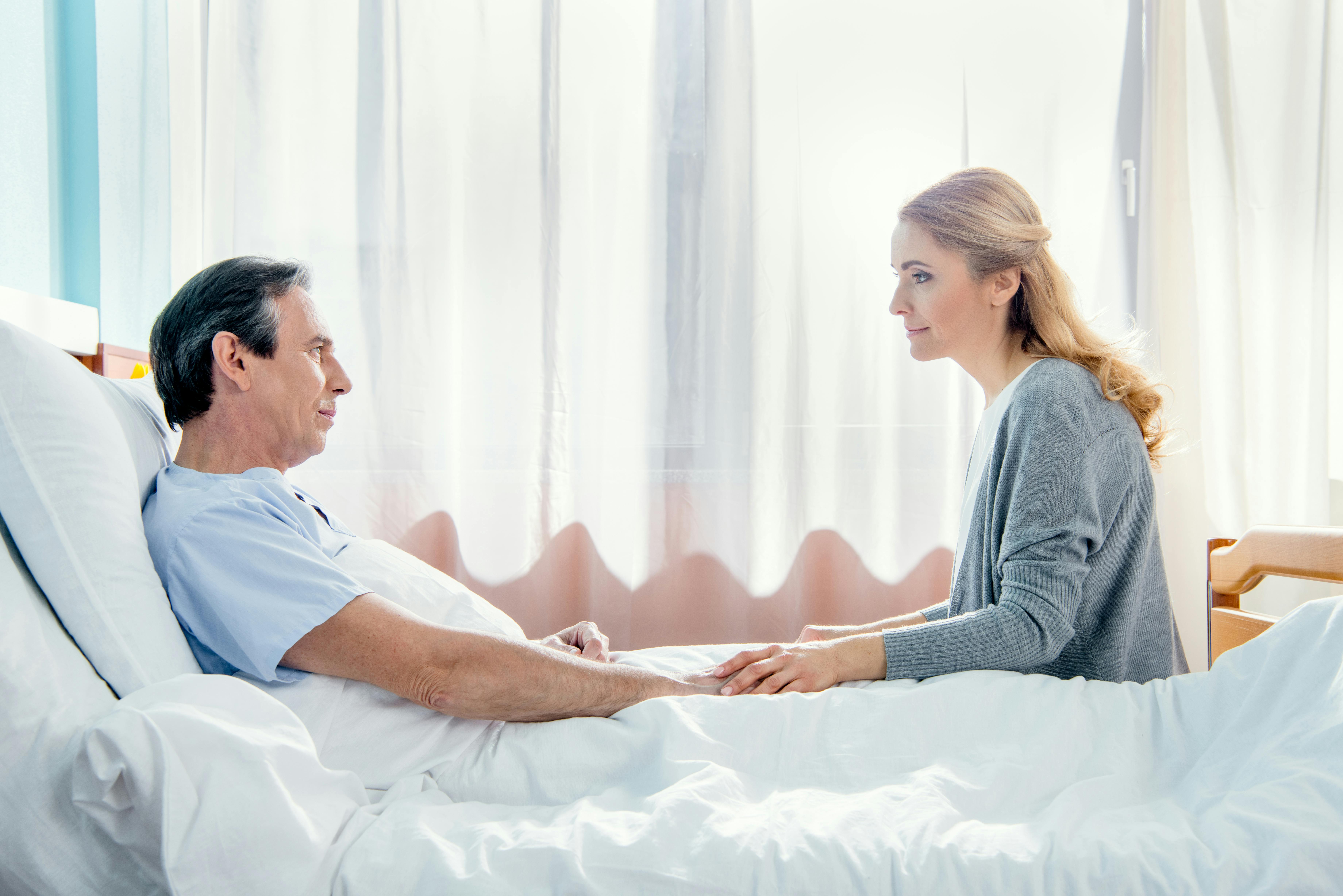 En kvinde sidder ved sin mands hospitalsseng. De holder hinanden i hånden og ser bekymrede ud.