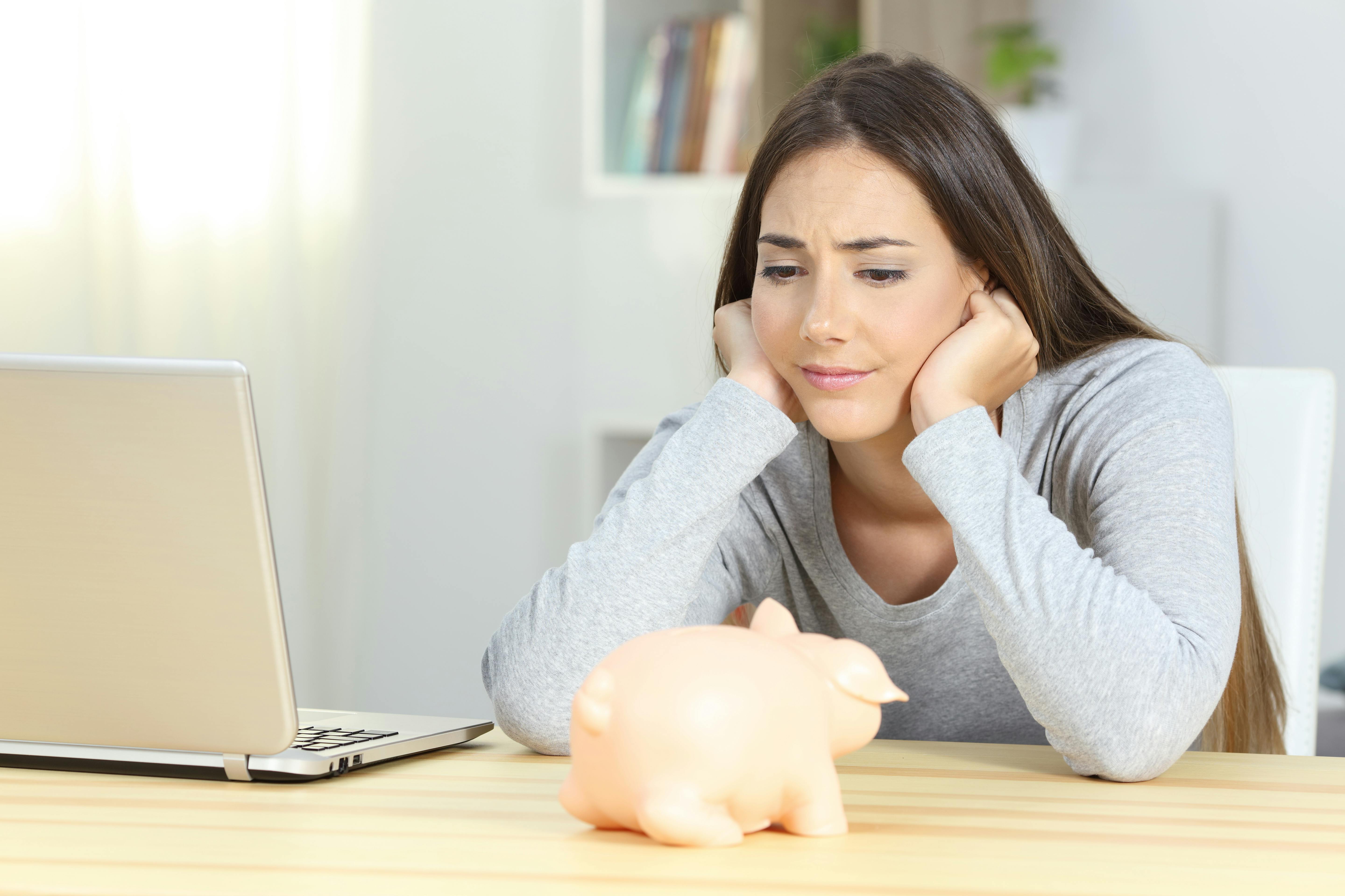 En ung kvinde sidder foran sin computer og en sparegris og ser opgivende ud.