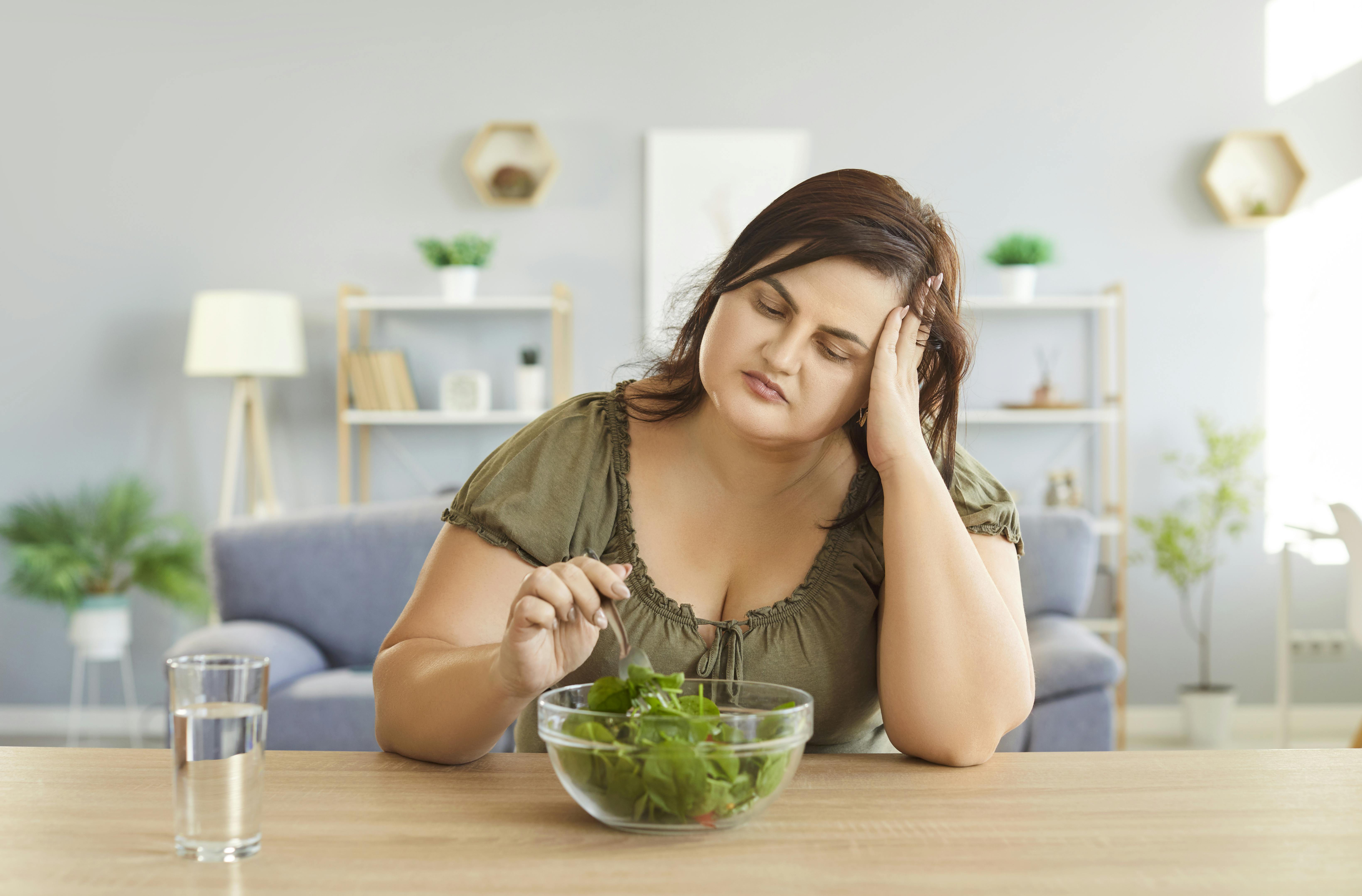 Overvægtig kvinde sidder og stikker til en skål med salat, mens hun tager sig opgivende til hovedet med den anden hånd.