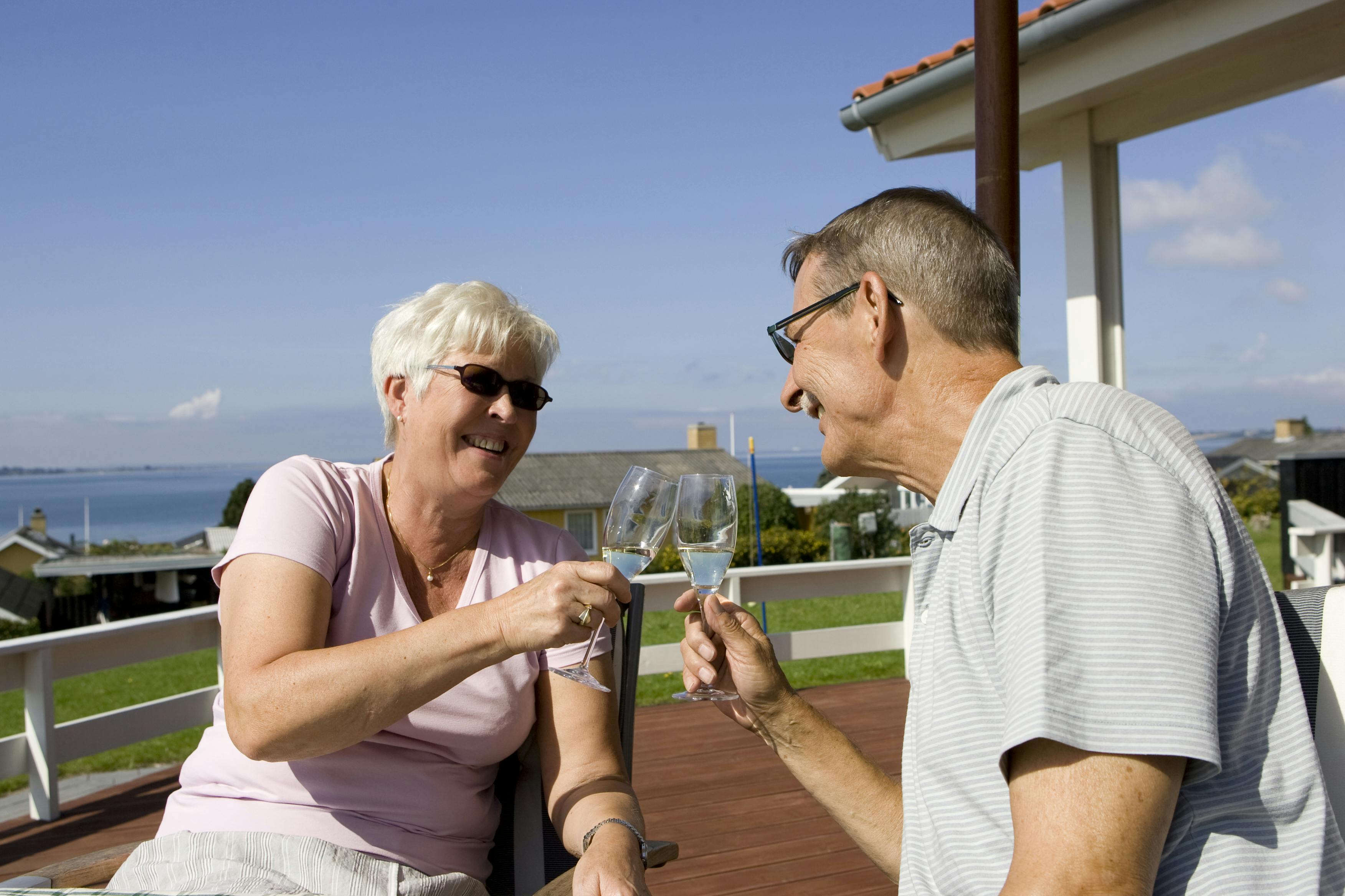 Et par pensionister skåler i champagne på en terrasse, hvor man ser havet i baggrunden.
