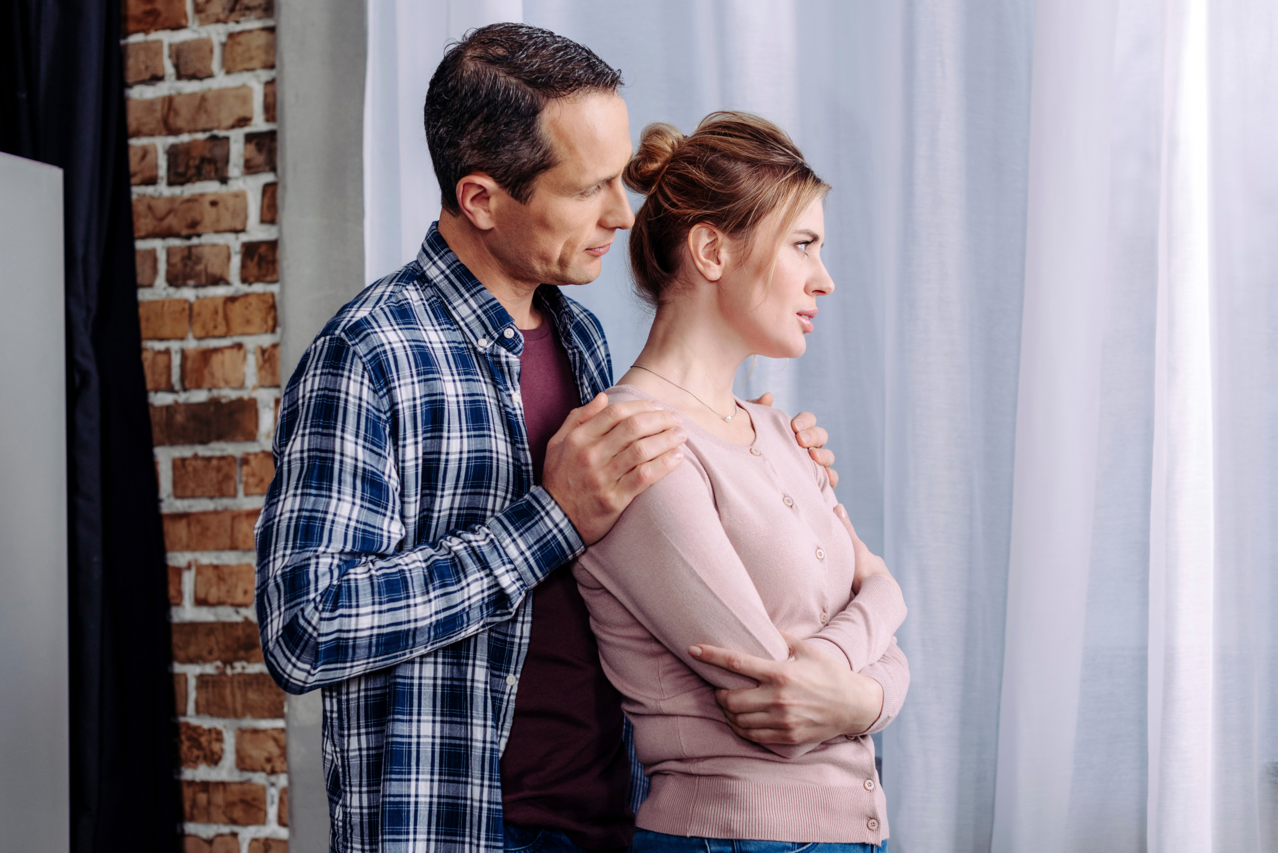 Kvinde står og ser længselsfuldt frem for sig, mens hendes mand står bag hende og holder på hendes skuldre.