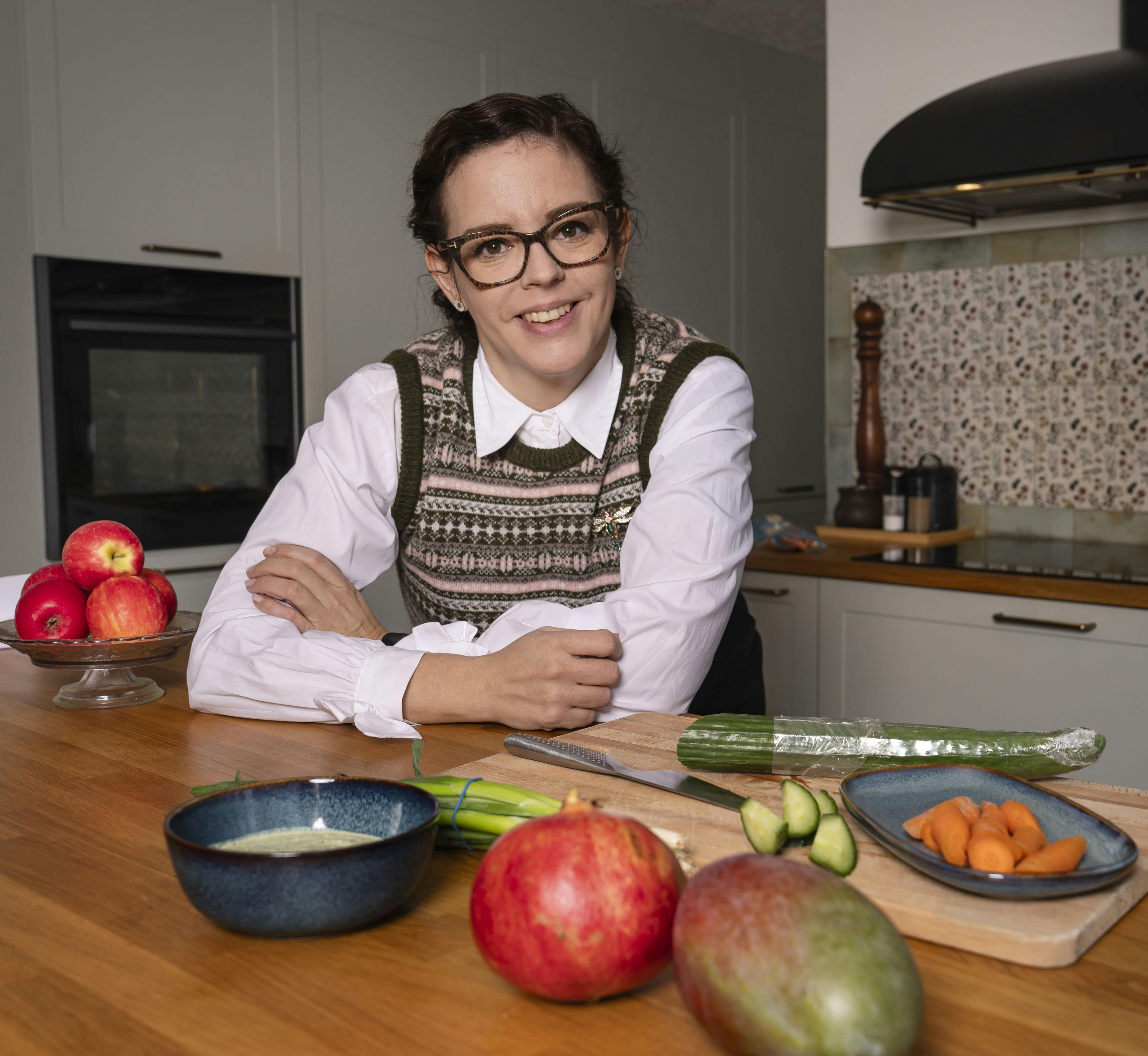 Tanja Madsen står i sit køkken med korslagte arme, som hun hviler på køkkenbordet. Hun har frugt og grønt, skærebræt og kniv liggende foran sig på køkkenbordet.