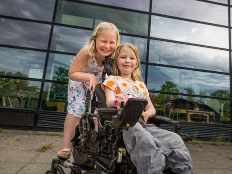 En lille pige står ved siden af sin bedste veninde, der sidder i kørestol