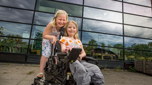 En lille pige står ved siden af sin bedste veninde, der sidder i kørestol