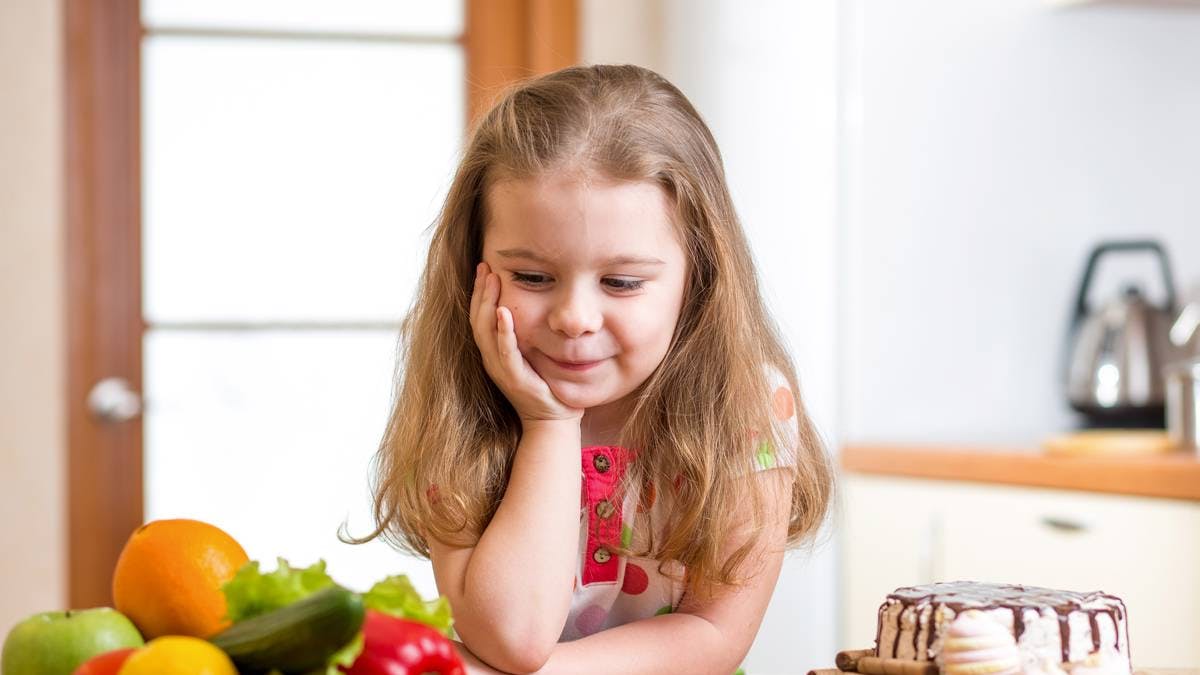 Lille piger kigger på fad med frugt, mens der står en fristende lagkage ved siden af hende.