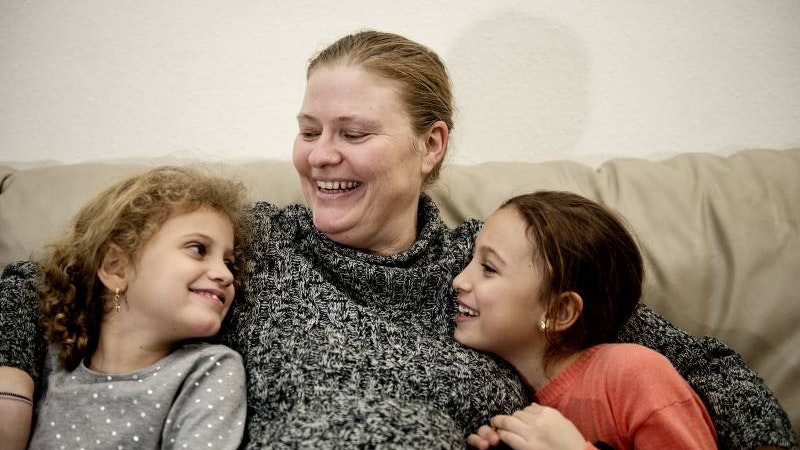 En mor og hendes to kønne døtre sidder sammen i en sofa og smiler til hinanden. Mor sidder med døtrene på hver sin side. De ser lykkelige ud